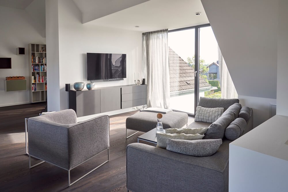 Heller Wohnbereich mit grauem Sofa von COR, minimalistischen Sesseln und modernem TV-Board in einer großzügigen Dachwohnung.